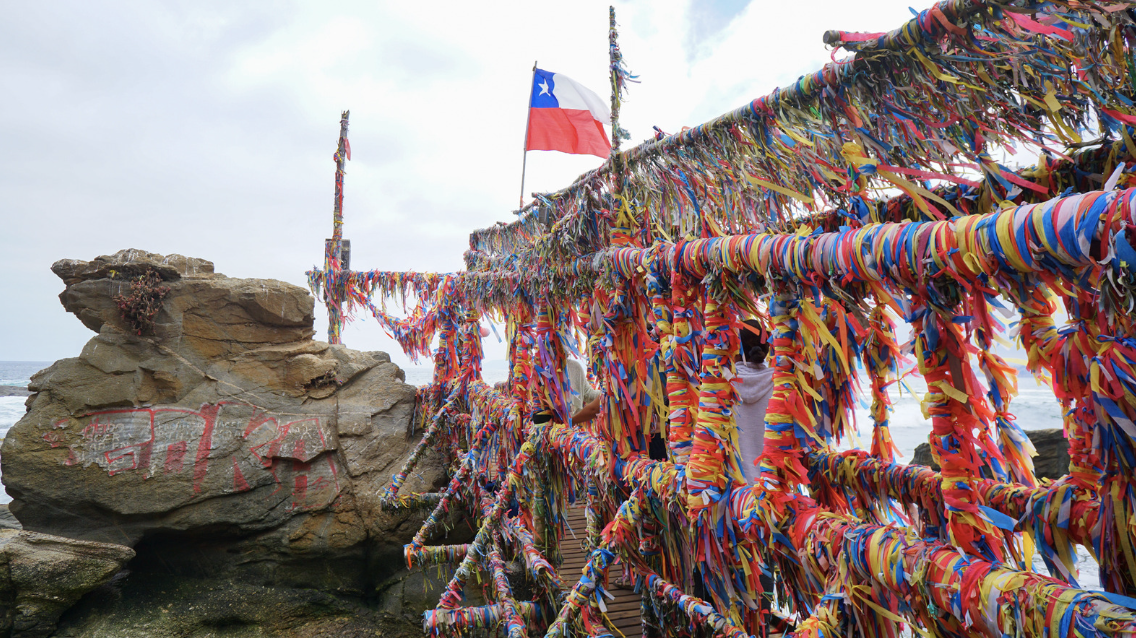 The railings of a bridge by the sea are covering in multi-colored ribbons. A Chilean flag is blowing in the wind in the background.